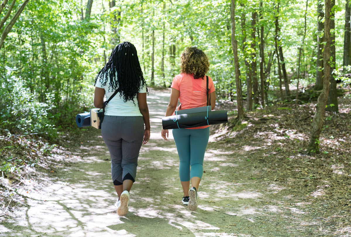 Two women carrying Mindtra benches and rolled yoga mats along a woodland path.